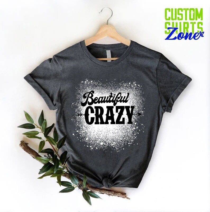 Puede incluir: Una camiseta gris oscuro con un dise&ntilde;o de pintura en aerosol blanca. El dise&ntilde;o presenta el texto "Beautiful CRAZY" en negro.