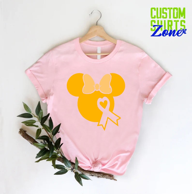 K&ouml;nnte beinhalten: Pinkes T-Shirt mit einer gelben Silhouette von Mickey Mouse mit Schleife und einem gelben Band mit einem Herz in der Mitte. Der Text "CUSTOM SHIRTS Zone" befindet sich in der oberen rechten Ecke des Bildes.