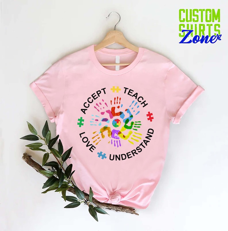 Puede incluir: Camiseta rosa con un dise&ntilde;o circular que presenta huellas de manos de colores y las palabras "Aceptar, Ense&ntilde;ar, Amar, Entender" alrededor del exterior. El dise&ntilde;o tambi&eacute;n incluye piezas de rompecabezas.
