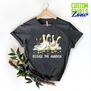 May include: A dark gray t-shirt with a graphic of four geese walking in a line. The text "Release the Quacken" is printed below the geese.