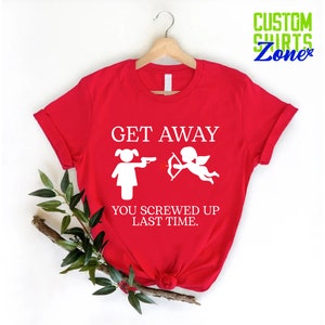 May include: Red t-shirt with a white graphic that says "Get Away You Screwed Up Last Time." The graphic features a stick figure of a woman holding a gun and a stick figure of Cupid holding a bow and arrow.