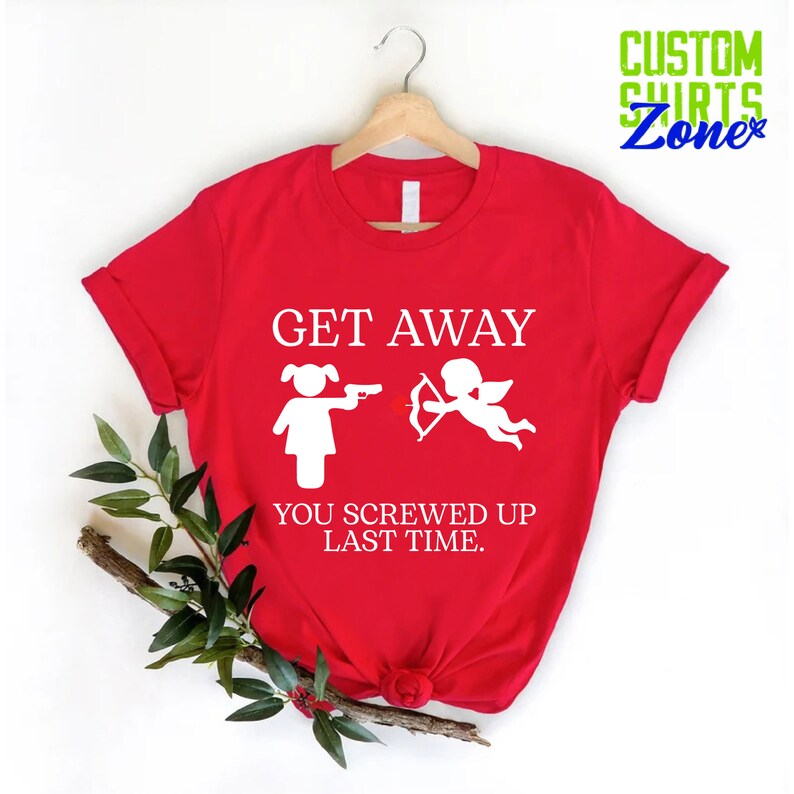 May include: Red t-shirt with a white graphic that says "Get Away You Screwed Up Last Time." The graphic features a stick figure of a woman holding a gun and a stick figure of Cupid holding a bow and arrow.