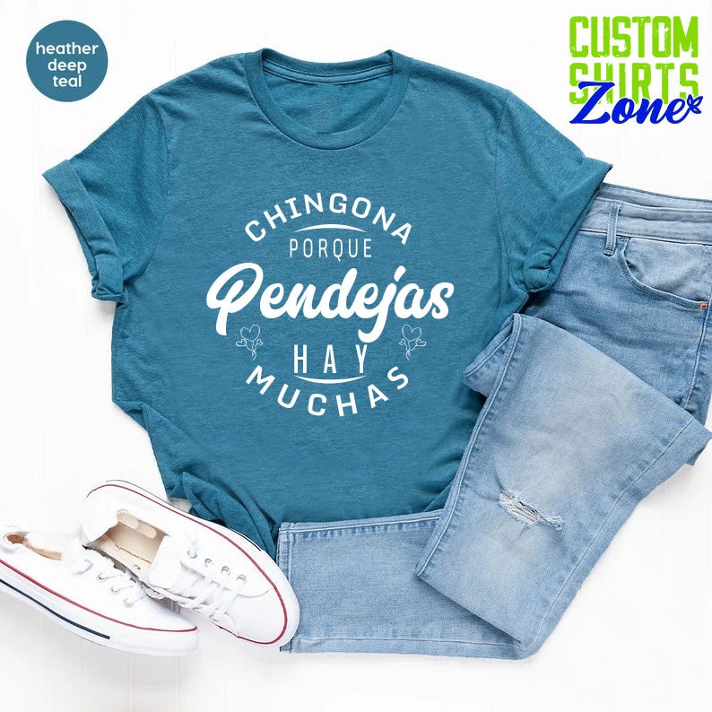 May include: A teal blue t-shirt with white text that reads "Chingona Porque Pendejas Hay Muchas". The shirt is laid flat on a white surface with a pair of white sneakers and blue jeans.