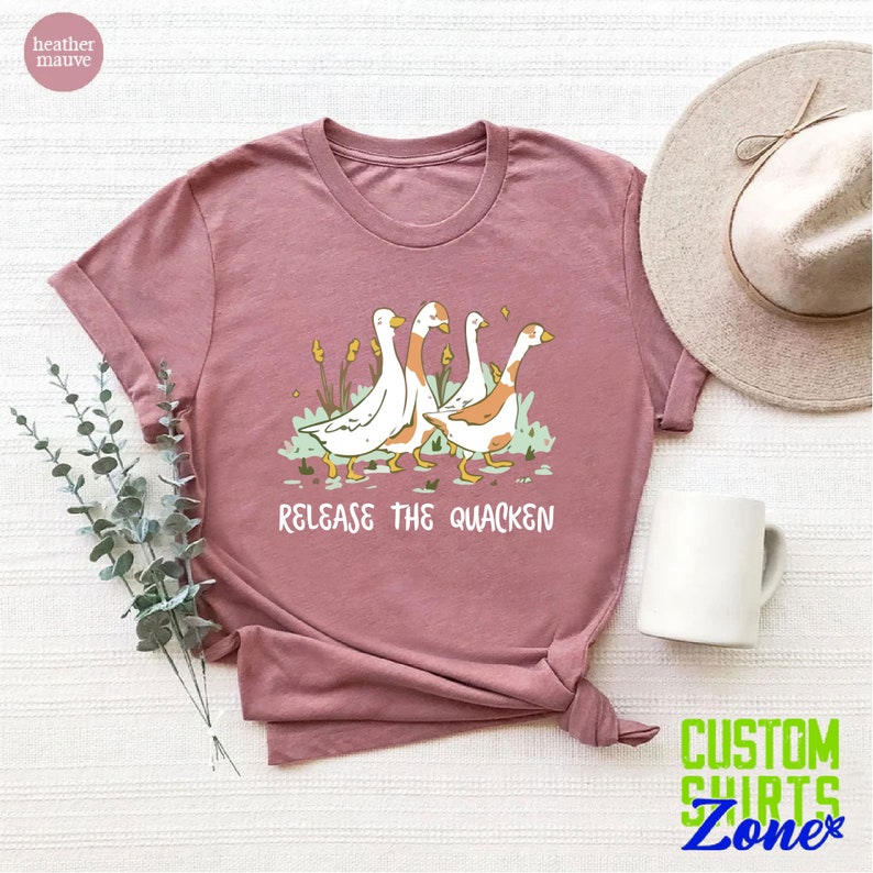 May include: A heather mauve t-shirt with a graphic of four geese walking in a line. The geese are white with orange accents. The text "Release the Quacken" is printed below the geese.