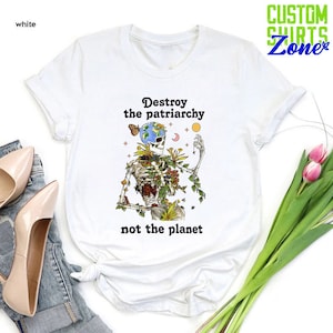 May include: White t-shirt with a graphic of a skeleton holding a globe with flowers and vines growing from its body. The text "Destroy the patriarchy not the planet" is printed below the graphic.