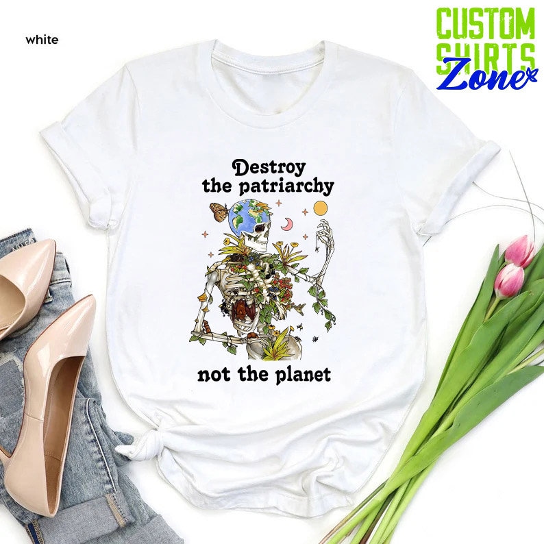 May include: White t-shirt with a graphic of a skeleton holding a globe with flowers and vines growing from its body. The text "Destroy the patriarchy not the planet" is printed below the graphic.