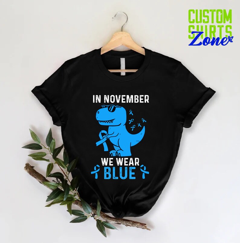 May include: Black t-shirt with a blue dinosaur graphic wearing sunglasses and holding a blue ribbon. The text reads "IN NOVEMBER WE WEAR BLUE" with blue ribbons. The shirt is hanging on a wooden hanger.