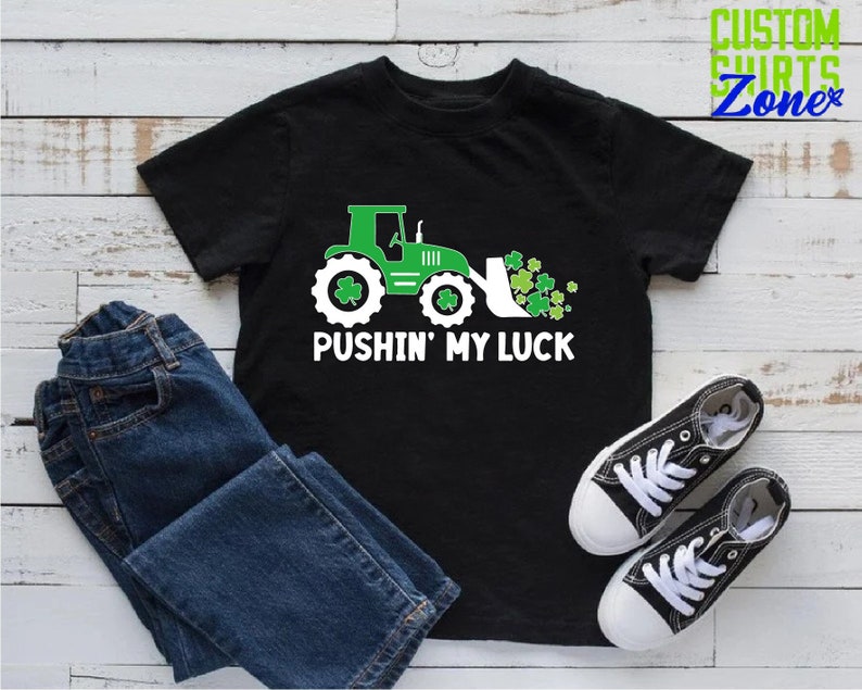 May include: A black t-shirt with a green tractor pushing a pile of shamrocks. The text "Pushin' My Luck" is printed in white below the tractor.