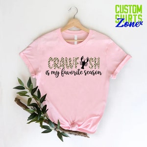 Puede incluir: Camiseta rosa con un dise&ntilde;o de estampado de leopardo negro y dorado que dice "Crawfish is my favorite season".