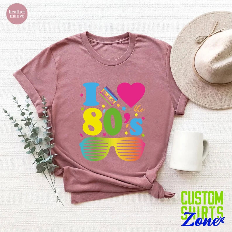 May include: A heather mauve t-shirt with a graphic design that says "I &hearts; the 80's" with a cassette tape, a pink heart, and rainbow sunglasses.