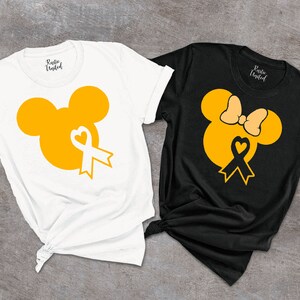 K&ouml;nnte beinhalten: Zwei T-Shirts mit einem gelben Mickey-Maus-Kopf-Design. Der Mickey-Maus-Kopf hat ein gelbes Band mit einem wei&szlig;en Herzen in der Mitte. Ein T-Shirt ist wei&szlig; und das andere ist schwarz. Beide T-Shirts haben den Text "Rustic United" auf der R&uuml;ckseite.