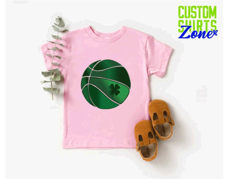 Puede incluir: Camiseta rosa con una pelota de baloncesto verde con un tr&eacute;bol. La camiseta dice "Custom Shirts Zone" en verde y azul.