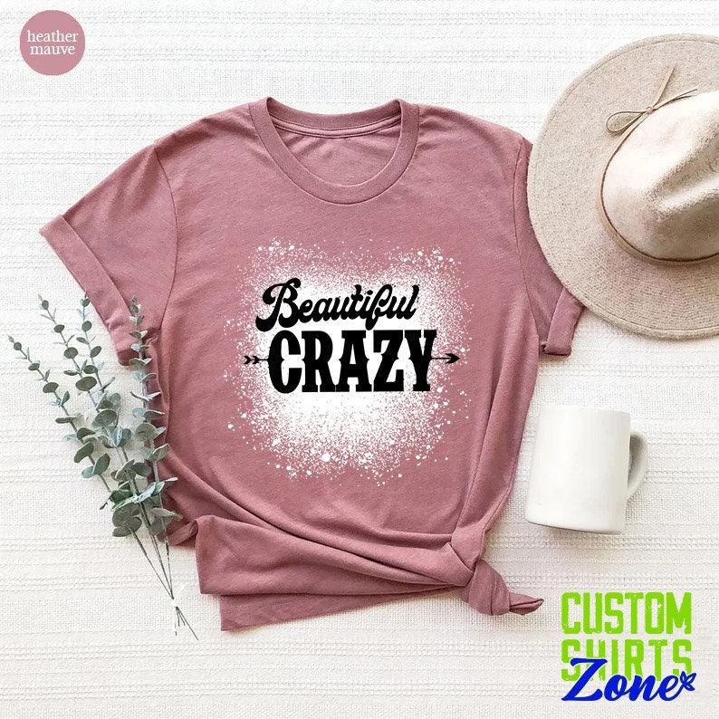 Puede incluir: Una camiseta de color malva con un dise&ntilde;o de salpicaduras blancas y el texto "Beautiful Crazy" en negro.