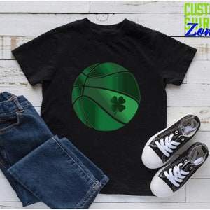 Puede incluir: Camiseta negra con una pelota de baloncesto verde con un tr&eacute;bol. La camiseta est&aacute; sobre una superficie de madera blanca con un par de zapatillas negras y blancas y unos vaqueros azules.