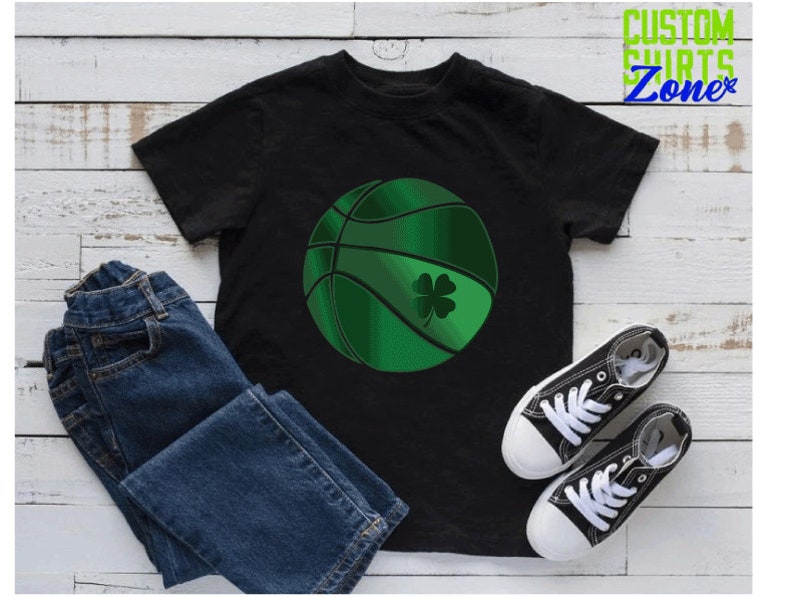Puede incluir: Camiseta negra con una pelota de baloncesto verde con un tr&eacute;bol. La camiseta est&aacute; sobre una superficie de madera blanca con un par de zapatillas negras y blancas y unos vaqueros azules.