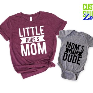 Op de afbeelding: Een bordeauxrood t-shirt met de witte tekst "LITTLE DUDE'S MOM", naast een grijze babyromper met de zwarte tekst "MOM'S LITTLE DUDE". De shirts zijn van Custom Shirts Zone.