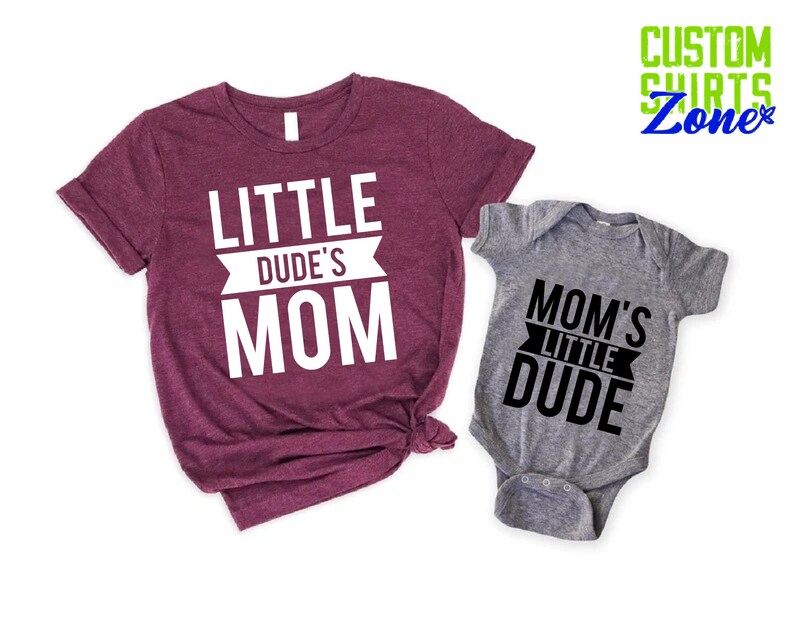 Op de afbeelding: Een bordeauxrood t-shirt met de witte tekst "LITTLE DUDE'S MOM", naast een grijze babyromper met de zwarte tekst "MOM'S LITTLE DUDE". De shirts zijn van Custom Shirts Zone.