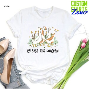 May include: White t-shirt with a graphic of four geese walking in a line. The geese are white with orange markings. The text "Release the Quacken" is printed below the geese.