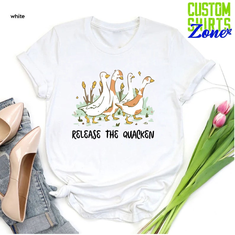 May include: White t-shirt with a graphic of four geese walking in a line. The geese are white with orange markings. The text "Release the Quacken" is printed below the geese.
