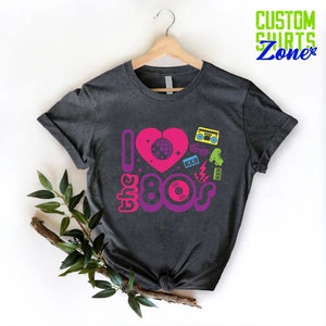 May include: A dark gray t-shirt with a pink heart and the text "I love the 80s" in purple and pink. The heart has a disco ball inside and is surrounded by 80s icons like a boombox, roller skates, sunglasses, and a cassette tape.