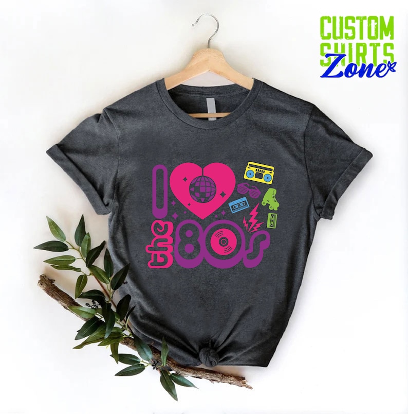 May include: A dark gray t-shirt with a pink heart and the text "I love the 80s" in purple and pink. The heart has a disco ball inside and is surrounded by 80s icons like a boombox, roller skates, sunglasses, and a cassette tape.