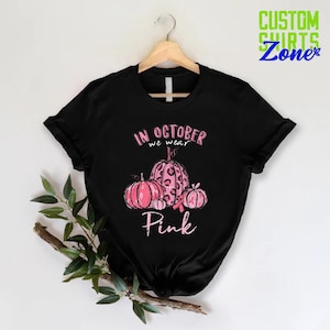 May include: Black t-shirt with a pink ribbon and pumpkins. The text on the shirt reads "In October we wear Pink."