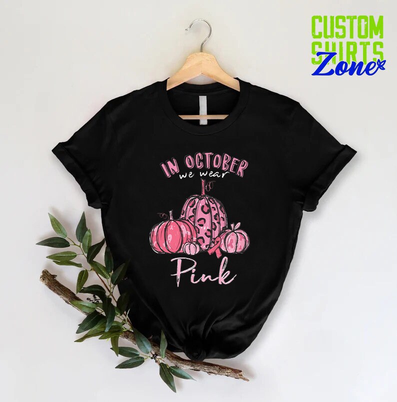 May include: Black t-shirt with a pink ribbon and pumpkins. The text on the shirt reads "In October we wear Pink."