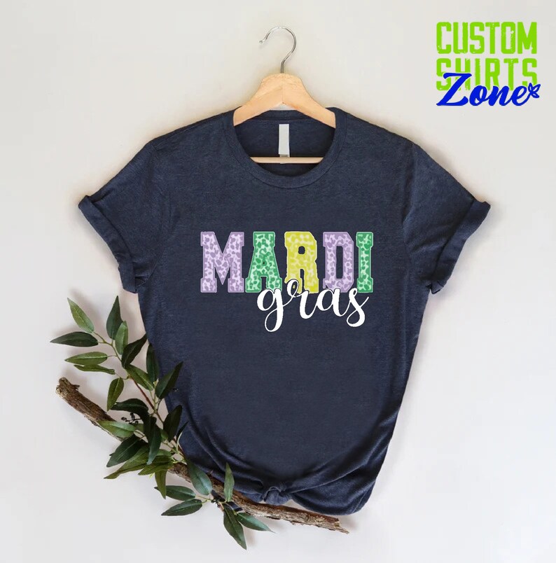 May include: A dark blue t-shirt with the words "Mardi Gras" printed on the front in purple, green, and yellow letters with a leopard print pattern.