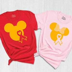K&ouml;nnte beinhalten: Zwei T-Shirts mit einem gelben Mickey Mouse und Minnie Mouse Design. Das Mickey Mouse Shirt ist rot und das Minnie Mouse Shirt ist rosa. Beide Shirts haben ein gelbes Band mit einem Herzen in der Mitte.
