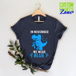 May include: Navy blue t-shirt with a blue dinosaur wearing sunglasses and holding a blue ribbon. The text on the shirt reads "IN NOVEMBER WE WEAR BLUE" and "CUSTOM SHIRTS Zone" in the upper right corner.