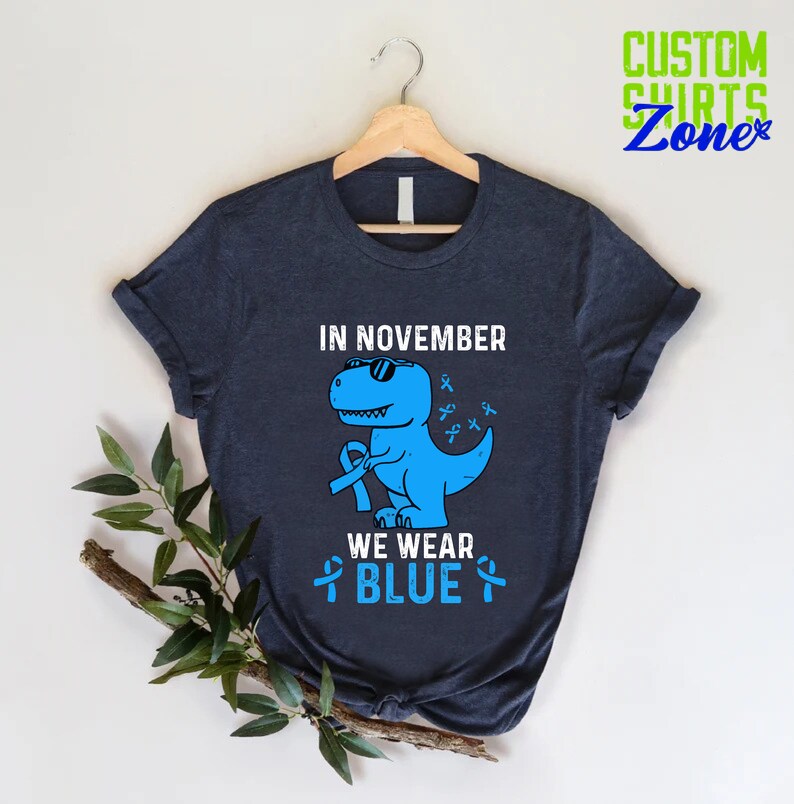 May include: Navy blue t-shirt with a blue dinosaur wearing sunglasses and holding a blue ribbon. The text on the shirt reads "IN NOVEMBER WE WEAR BLUE" and "CUSTOM SHIRTS Zone" in the upper right corner.