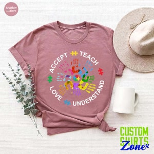 Puede incluir: Una camiseta de color malva con un dise&ntilde;o circular blanco con las palabras "Aceptar, Amar, Ense&ntilde;ar, Entender" y huellas de manos de colores alrededor de una pieza de rompecabezas.