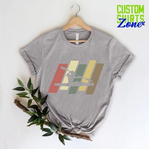 K&ouml;nnte beinhalten: Graues T-Shirt mit einer Retro-Grafik einer Person im Kajak. Die Grafik besteht aus f&uuml;nf vertikalen Streifen in Rot-, Gelb-, Gr&uuml;n- und Braunt&ouml;nen.