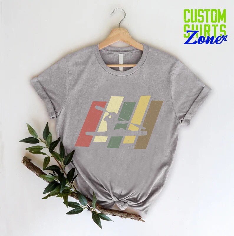 K&ouml;nnte beinhalten: Graues T-Shirt mit einer Retro-Grafik einer Person im Kajak. Die Grafik besteht aus f&uuml;nf vertikalen Streifen in Rot-, Gelb-, Gr&uuml;n- und Braunt&ouml;nen.