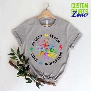 Puede incluir: Camiseta gris con un dise&ntilde;o circular que presenta huellas de manos de colores y las palabras "Aceptar", "Ense&ntilde;ar", "Amar" y "Entender".