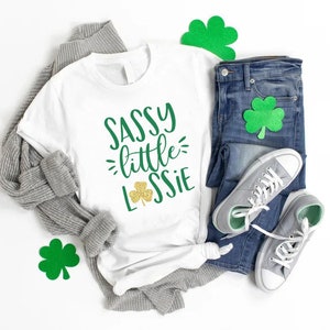 K&ouml;nnte beinhalten: Wei&szlig;es T-Shirt mit dem Text "Sassy little Lassie" in gr&uuml;ner und goldener Glitzer-Schrift. Ein Kleeblatt ist im Design enthalten. Das Shirt liegt auf einem wei&szlig;en Hintergrund mit einem grauen Pullover, einer blauen Jeans und gr&uuml;nen Kleeblatt-Ausschnitten.