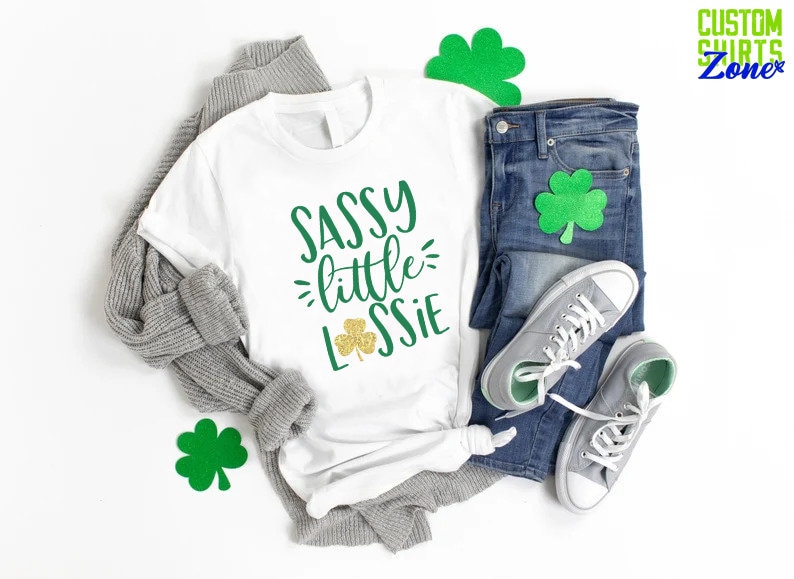 K&ouml;nnte beinhalten: Wei&szlig;es T-Shirt mit dem Text "Sassy little Lassie" in gr&uuml;ner und goldener Glitzer-Schrift. Ein Kleeblatt ist im Design enthalten. Das Shirt liegt auf einem wei&szlig;en Hintergrund mit einem grauen Pullover, einer blauen Jeans und gr&uuml;nen Kleeblatt-Ausschnitten.