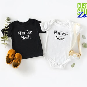 May include: Two baby shirts, one black and one white, with the text "N is for Noah" printed on them. The shirts are laid out on a white surface.