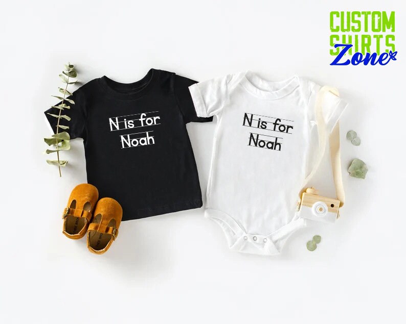 May include: Two baby shirts, one black and one white, with the text "N is for Noah" printed on them. The shirts are laid out on a white surface.