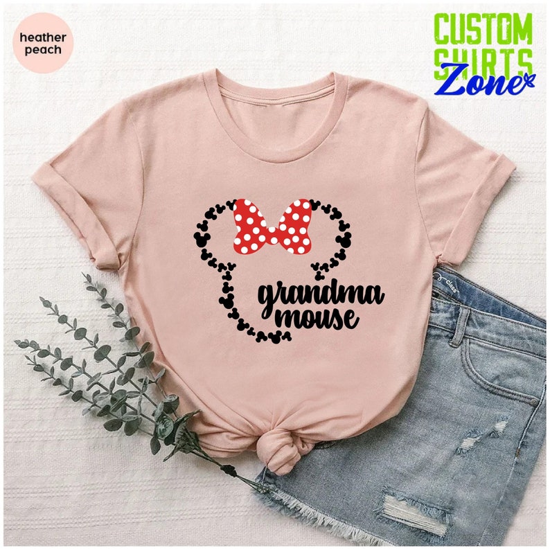 Op de afbeelding: Perzikroze t-shirt met een Minnie Mouse-ontwerp. Het ontwerp bevat een rode strik met witte stippen en de zwarte tekst "grandma mouse". Het t-shirt heeft korte mouwen en is gemaakt van zacht materiaal.