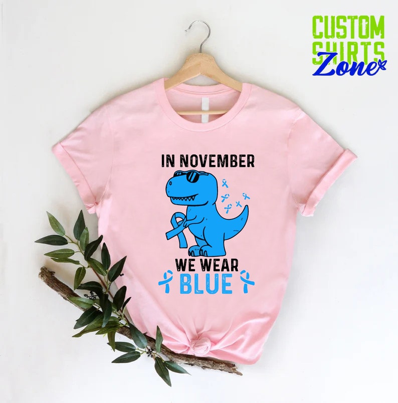 May include: A light pink t-shirt with the text "IN NOVEMBER WE WEAR BLUE". The shirt features a blue dinosaur wearing sunglasses and holding a blue ribbon. Additional blue ribbons are scattered around the dinosaur.