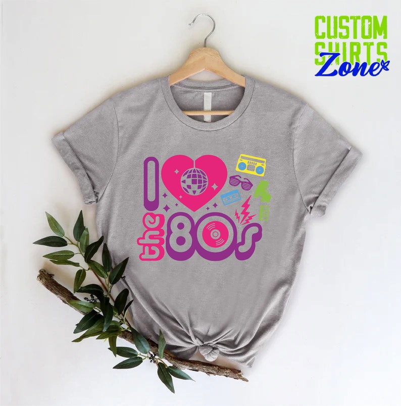 May include: A gray t-shirt with a graphic design that says "I love the 80s" with a pink heart and 80s icons like a boombox, sunglasses, cassette tape, lightning bolt, and a record.