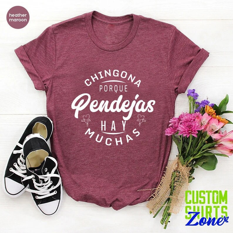 May include: A heather maroon t-shirt with the text "Chingona Porque Pendejas Hay Muchas" printed in white. The shirt is laid flat on a white surface with a bouquet of flowers and a pair of black sneakers.
