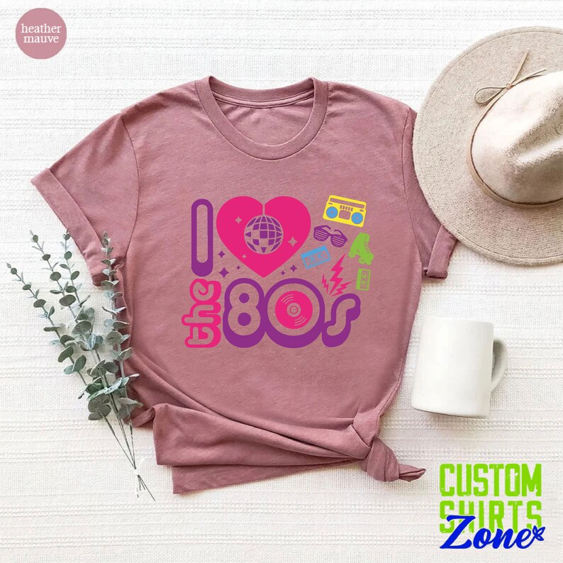 May include: A heather mauve t-shirt with a graphic design that says "I love the 80s" with a pink heart and 80s icons like a disco ball, boombox, sunglasses, and a record.