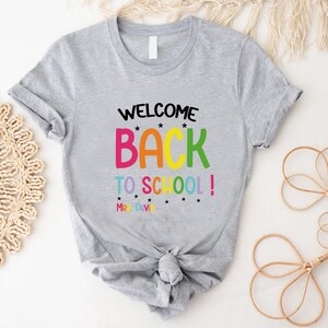 以下が含まれることがあります： カラフルな文字で「WELCOME BACK TO SCHOOL！」と書かれたライトグレーのTシャツ。テキストは小さな黒い星でアクセントがつけられています。シャツはウエストで結ばれています。