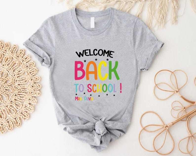 以下が含まれることがあります： カラフルな文字で「WELCOME BACK TO SCHOOL！」と書かれたライトグレーのTシャツ。テキストは小さな黒い星でアクセントがつけられています。シャツはウエストで結ばれています。