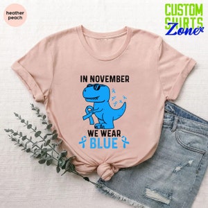 May include: Heather peach t-shirt featuring a blue dinosaur wearing sunglasses and holding a blue ribbon. The text "IN NOVEMBER WE WEAR BLUE" is printed above and below the dinosaur. The shirt is folded and styled with denim shorts.