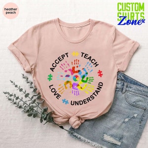 Puede incluir: Una camiseta rosa con un dise&ntilde;o circular que presenta huellas de manos coloridas y piezas de rompecabezas. El texto alrededor del c&iacute;rculo dice "Aceptar, Ense&ntilde;ar, Amar, Entender".