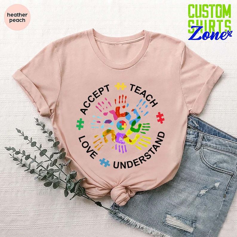 Puede incluir: Una camiseta rosa con un dise&ntilde;o circular que presenta huellas de manos coloridas y piezas de rompecabezas. El texto alrededor del c&iacute;rculo dice "Aceptar, Ense&ntilde;ar, Amar, Entender".