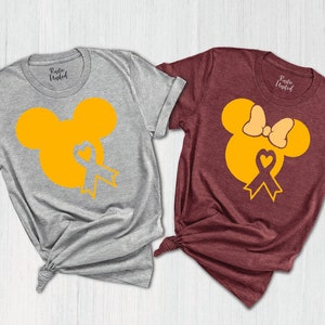 K&ouml;nnte beinhalten: Zwei T-Shirts mit einem gelben Mickey Mouse und Minnie Mouse Design. Das Mickey Mouse Shirt ist grau und das Minnie Mouse Shirt ist weinrot. Beide Shirts haben ein gelbes Band mit einem Herzen in der Mitte.
